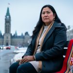 Indigenous woman in winter clothing with a Métis-patterned sash-like textile in the foreground, with a softly blurred Parliament Hill silhouette in the background.